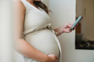 Side view crop unrecognizable pregnant woman text messaging on phone while standing in room in daylight